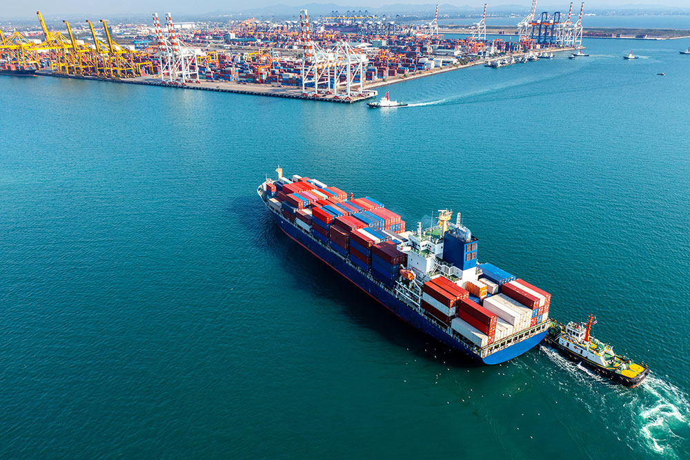 Aerial view of a large cargo ship laden with containers, approaching a bustling port with cranes and infrastructure, set against the clear blue sea.