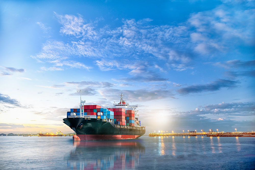 A large cargo ship loaded with colorful containers, docked at a port during sunset, with a vibrant sky and calm water reflecting the ship's silhouette.